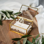 Hochwertige, personalisierbare Ringbox aus Glas mit goldfarbenem Metallrahmen, geöffnet und mit weißer Vinylschrift auf dem Deckel sowie einem aufgedruckten Hochzeitsdatum auf der Vorderseite. Im Inneren liegt ein beiges Ringkissen mit zarter Schleife, dekoriert mit kleinen weißen Blüten. Die Box befindet sich auf einer Holzoberfläche, umgeben von weichen, weißen Stoffen und Olivenzweigen im natürlichen Vintage-Stil, ideal für Hochzeit, Verlobung und Eheringe.