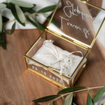 Hochwertige, personalisierbare Ringbox aus Glas mit goldfarbenem Metallrahmen, geöffnet und mit weißer Vinylschrift auf dem Deckel sowie einem aufgedruckten Hochzeitsdatum auf der Vorderseite. Im Inneren liegt ein beiges Ringkissen mit zarter Schleife, dekoriert mit kleinen weißen Blüten. Die Box befindet sich auf einer Holzoberfläche, umgeben von weichen, weißen Stoffen und Olivenzweigen im natürlichen Vintage-Stil, ideal für Hochzeit, Verlobung und Eheringe.