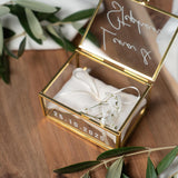 Hochwertige, personalisierbare Ringbox aus Glas mit goldfarbenem Metallrahmen, geöffnet und mit weißer Vinylschrift auf dem Deckel sowie einem aufgedruckten Hochzeitsdatum auf der Vorderseite. Im Inneren liegt ein beiges Ringkissen mit zarter Schleife, dekoriert mit kleinen weißen Blüten. Die Box befindet sich auf einer Holzoberfläche, umgeben von weichen, weißen Stoffen und Olivenzweigen im natürlichen Vintage-Stil, ideal für Hochzeit, Verlobung und Eheringe.