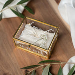 Hochwertige, personalisierbare Ringbox aus Glas mit goldfarbenem Metallrahmen, geöffnet und mit weißer Vinylschrift auf dem Deckel sowie einem aufgedruckten Hochzeitsdatum auf der Vorderseite. Im Inneren liegt ein beiges Ringkissen mit zarter Schleife, dekoriert mit kleinen weißen Blüten. Die Box befindet sich auf einer Holzoberfläche, umgeben von weichen, weißen Stoffen und Olivenzweigen im natürlichen Vintage-Stil, ideal für Hochzeit, Verlobung und Eheringe.