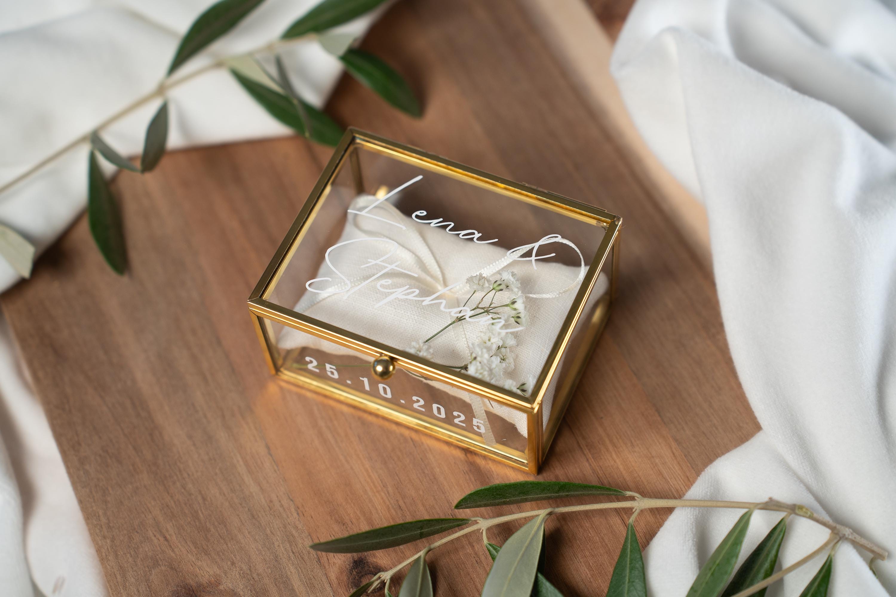 Hochwertige, personalisierbare Ringbox aus Glas mit goldfarbenem Metallrahmen, geöffnet und mit weißer Vinylschrift auf dem Deckel sowie einem aufgedruckten Hochzeitsdatum auf der Vorderseite. Im Inneren liegt ein beiges Ringkissen mit zarter Schleife, dekoriert mit kleinen weißen Blüten. Die Box befindet sich auf einer Holzoberfläche, umgeben von weichen, weißen Stoffen und Olivenzweigen im natürlichen Vintage-Stil, ideal für Hochzeit, Verlobung und Eheringe.